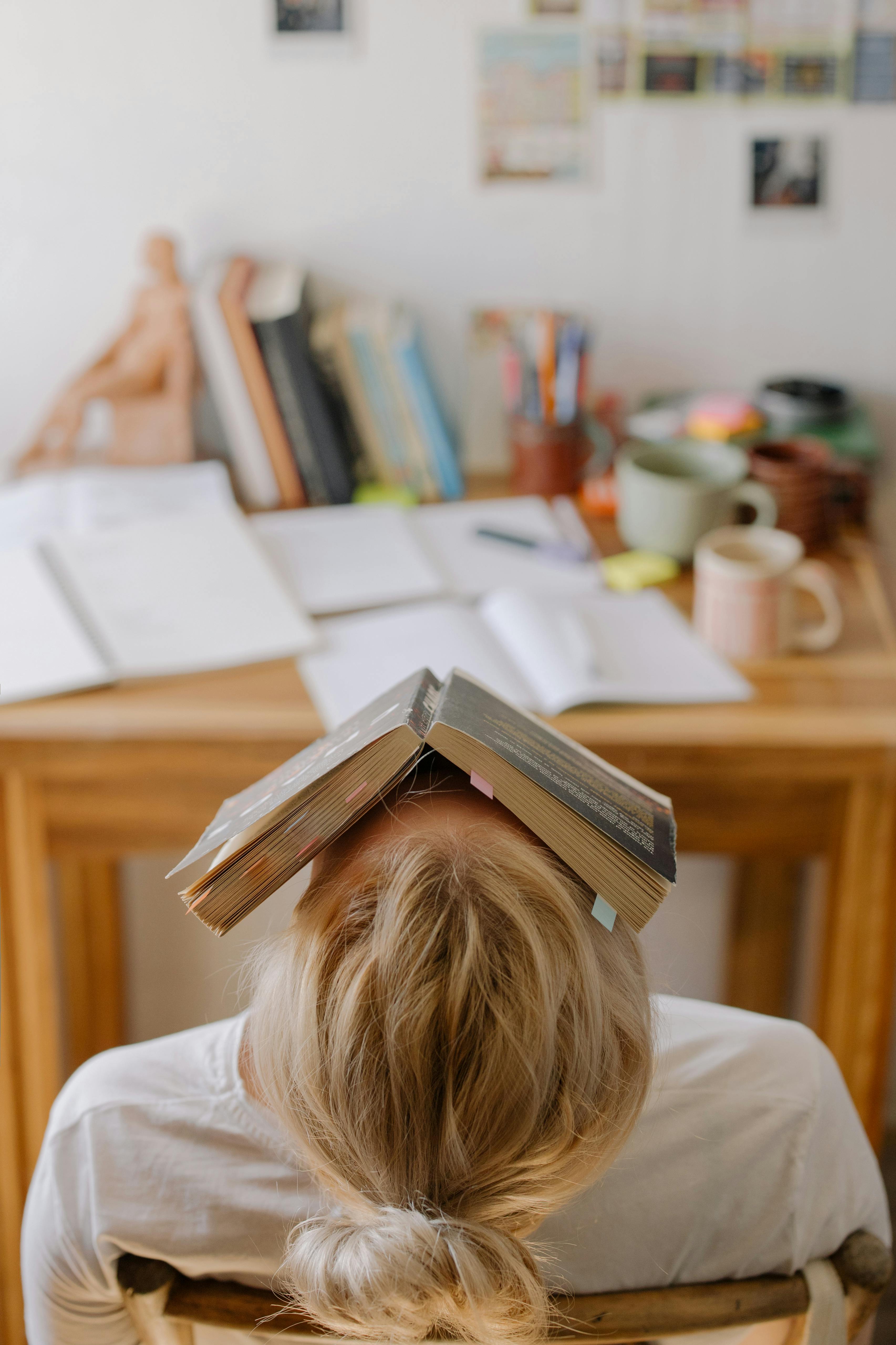 Student feeling stress and exhaustion while studying at a cluttered desk with an open book on their head.