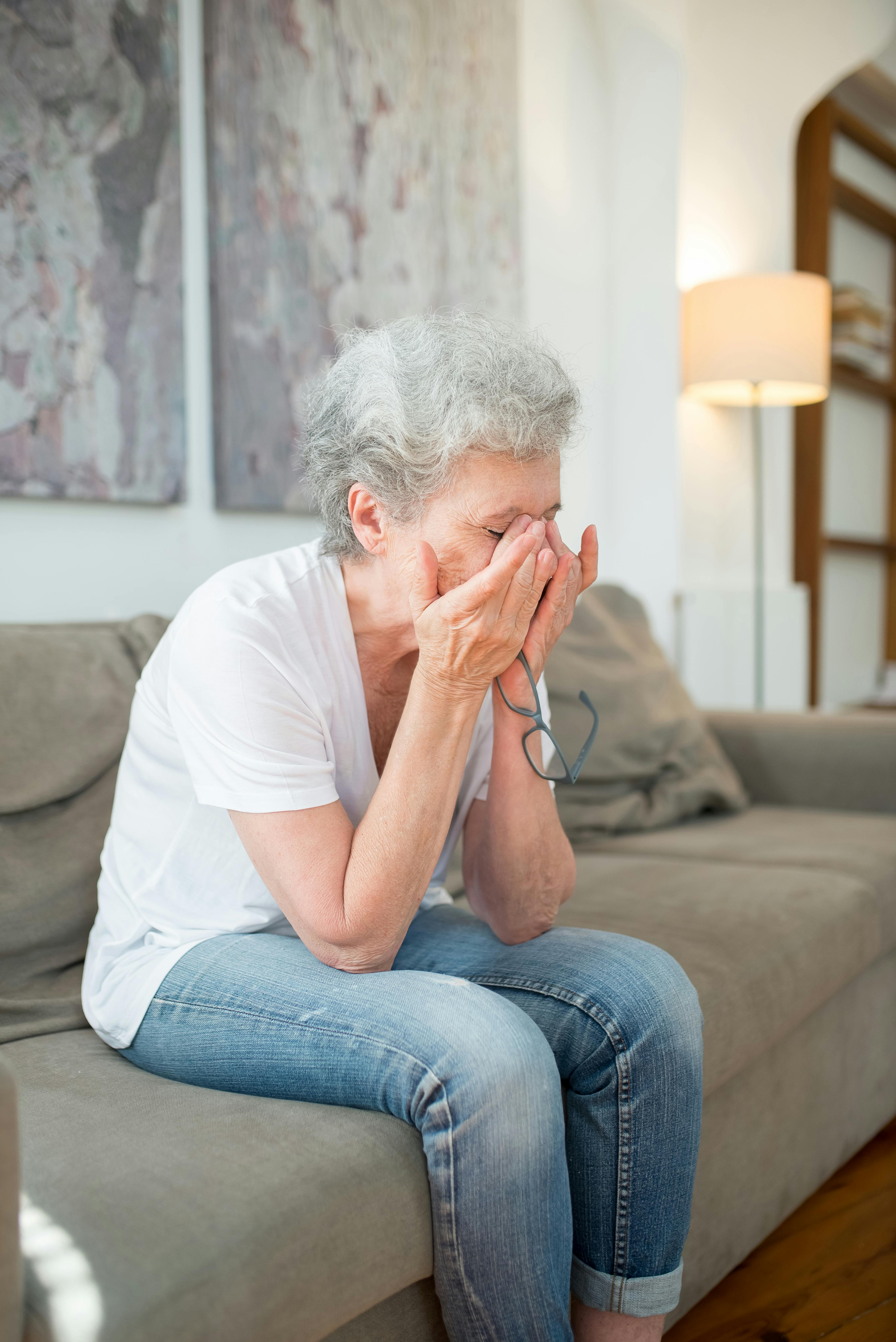 Senior woman with gray hair showing grief and emotion while sitting on a sofa indoors.